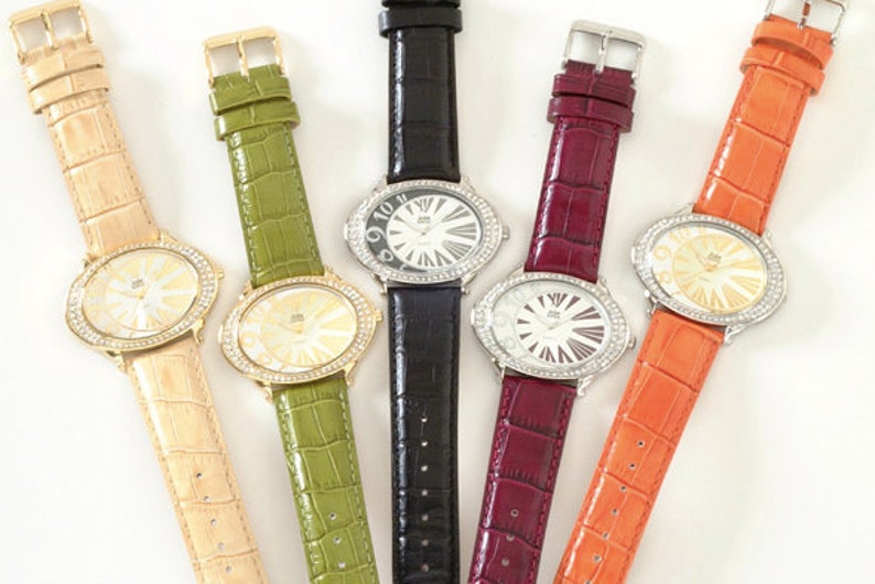 May include: Five oval-faced watches with rhinestone bezels and textured leather straps in beige, green, black, burgundy, and orange. The watch faces have a sunburst design and the straps have gold or silver buckles.