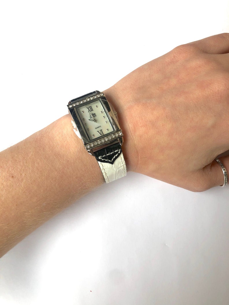 Puede incluir: Un reloj rectangular plateado con esfera blanca y n&uacute;meros romanos. El reloj tiene una correa con estampado en blanco y negro y est&aacute; adornado con peque&ntilde;os cristales transparentes alrededor de la esfera.
