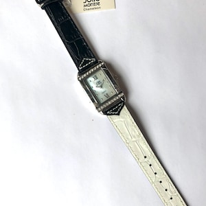 Puede incluir: Un reloj Jolie Montre Chameleon rectangular con esfera blanca, n&uacute;meros romanos y bisel plateado adornado con peque&ntilde;os cristales. La correa es de cuero negro que cambia a cuero blanco. Una etiqueta con el nombre de la marca est&aacute; adjunta.