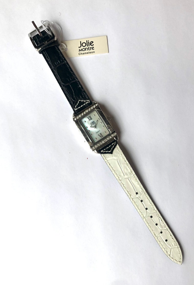 Puede incluir: Un reloj Jolie Montre Chameleon rectangular con esfera blanca, n&uacute;meros romanos y bisel plateado adornado con peque&ntilde;os cristales. La correa es de cuero negro que cambia a cuero blanco. Una etiqueta con el nombre de la marca est&aacute; adjunta.
