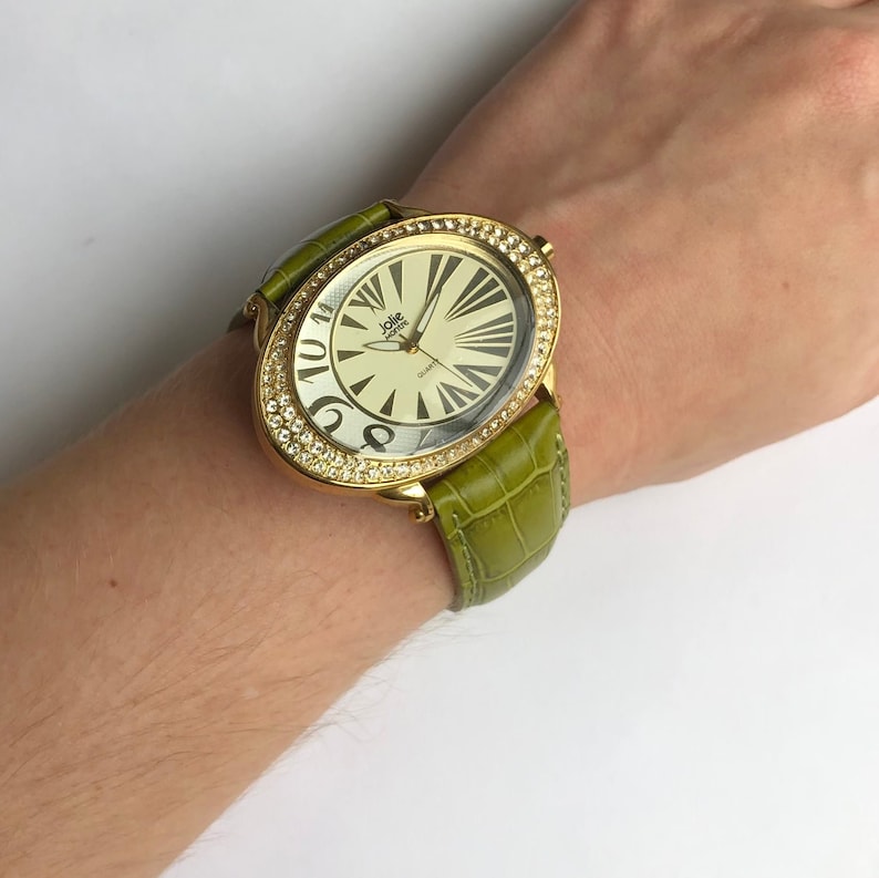 May include: Gold-toned oval watch with a cream-colored face and black numerals. The watch is adorned with a ring of clear crystals and a green leather band. The watch face reads "Jolie Moore" and "Quartz".