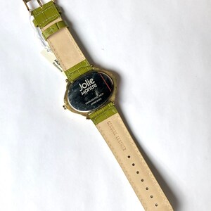 May include: A Jolie Montre watch with a light green leather band and gold-colored buckle. The watch face is oval-shaped with the brand name and "Stainless Steel Back Japan Movt" printed on it. The band is approximately 9 inches long.