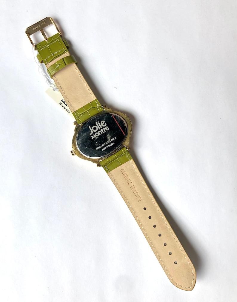 May include: A Jolie Montre watch with a light green leather band and gold-colored buckle. The watch face is oval-shaped with the brand name and "Stainless Steel Back Japan Movt" printed on it. The band is approximately 9 inches long.
