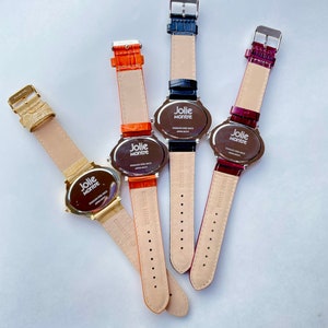 May include: Four Jolie Montre watches with leather straps in gold, orange, navy blue, and burgundy. The watch faces are oval-shaped with the brand name printed on them. The watch straps are in various colors.