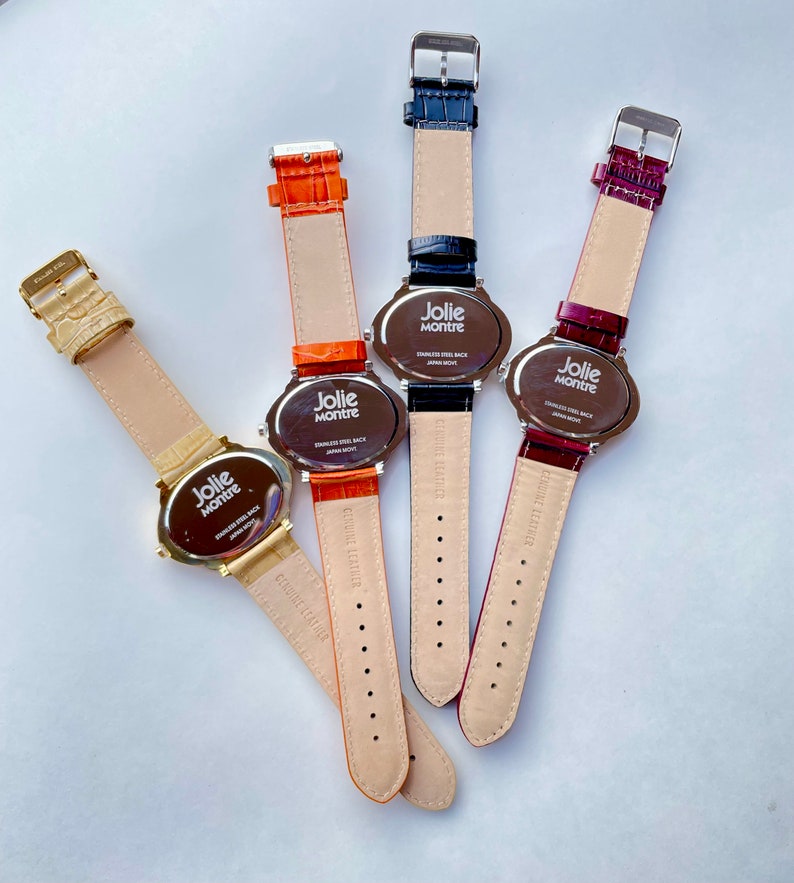 May include: Four Jolie Montre watches with leather straps in gold, orange, navy blue, and burgundy. The watch faces are oval-shaped with the brand name printed on them. The watch straps are in various colors.