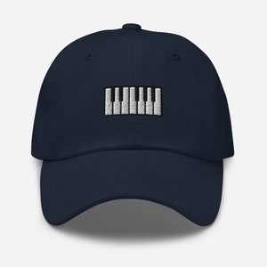 May include: Navy blue baseball cap featuring an embroidered piano keyboard design. The cap has a curved brim and an adjustable strap at the back. The piano keys are white and black, providing a striking contrast against the dark blue fabric.