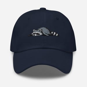 K&ouml;nnte beinhalten: Marineblaue Baseballkappe mit einem gestickten Waschb&auml;ren-Design. Der Waschb&auml;r ist in Grau- und Wei&szlig;t&ouml;nen dargestellt und liegt. Die Kappe hat einen gebogenen Schirm und einen Knopf oben.