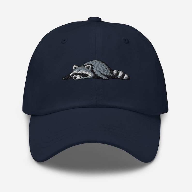 K&ouml;nnte beinhalten: Marineblaue Baseballkappe mit einem gestickten Waschb&auml;ren-Design. Der Waschb&auml;r ist in Grau- und Wei&szlig;t&ouml;nen dargestellt und liegt. Die Kappe hat einen gebogenen Schirm und einen Knopf oben.