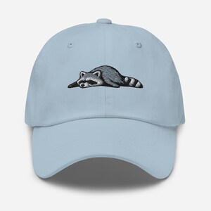 K&ouml;nnte beinhalten: Hellblaue Baseballkappe mit einem gestickten Waschb&auml;ren-Design. Der Waschb&auml;r ist in Grau- und Schwarzt&ouml;nen dargestellt und liegt. Die Kappe hat einen gebogenen Schirm und einen verstellbaren Riemen.