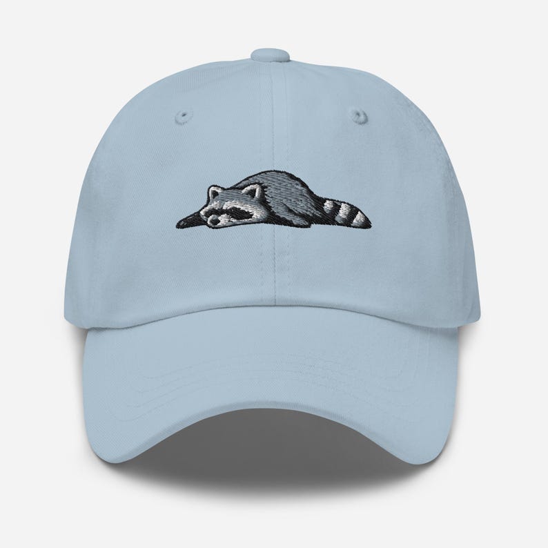 K&ouml;nnte beinhalten: Hellblaue Baseballkappe mit einem gestickten Waschb&auml;ren-Design. Der Waschb&auml;r ist in Grau- und Schwarzt&ouml;nen dargestellt und liegt. Die Kappe hat einen gebogenen Schirm und einen verstellbaren Riemen.