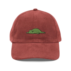 Könnte beinhalten: Ziegelrote Cord-Baseballkappe mit einem grünen, gestickten Froschdesign. Der Frosch liegt auf der Vorderseite der Kappe. Die Kappe hat einen gebogenen Schirm und einen verstellbaren Riemen auf der Rückseite.