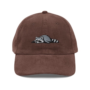 K&ouml;nnte beinhalten: Braune Cord-Baseballkappe mit einem gestickten Waschb&auml;ren-Design. Der Waschb&auml;r ist in Grau und Wei&szlig; dargestellt und liegt. Die Kappe hat einen gebogenen Schirm und einen verstellbaren Riemen am R&uuml;cken.