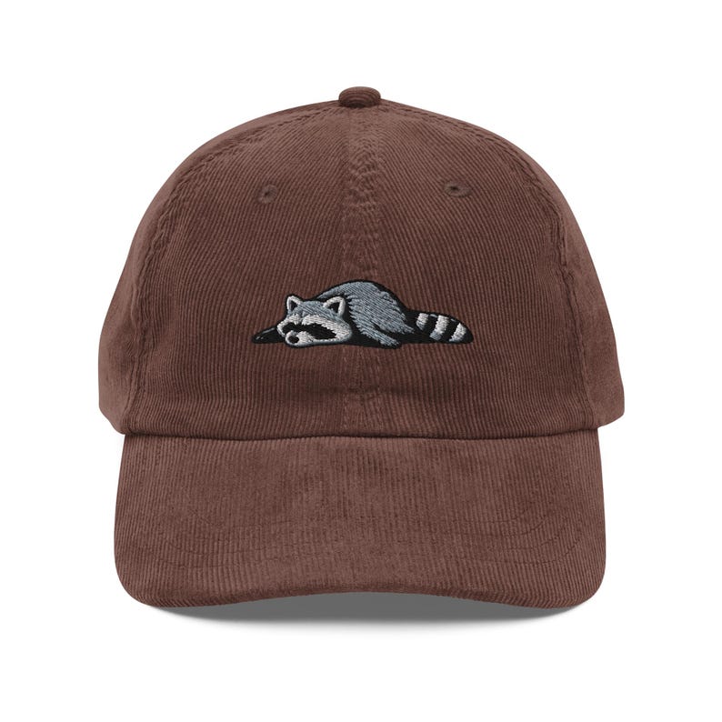 K&ouml;nnte beinhalten: Braune Cord-Baseballkappe mit einem gestickten Waschb&auml;ren-Design. Der Waschb&auml;r ist in Grau und Wei&szlig; dargestellt und liegt. Die Kappe hat einen gebogenen Schirm und einen verstellbaren Riemen am R&uuml;cken.