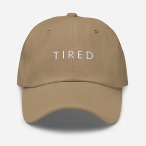 May include: A khaki dad hat with white embroidered text that reads "TIRED".