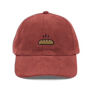 May include: A red corduroy baseball cap with an embroidered image of a brown baguette with steam coming off of it.
