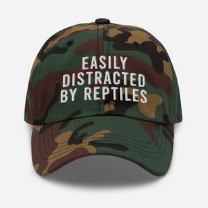 May include: A camouflage baseball cap with the text "EASILY DISTRACTED BY REPTILES" embroidered in white.