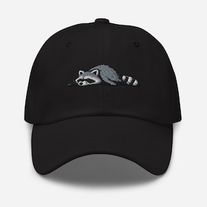 K&ouml;nnte beinhalten: Schwarze Baseballkappe mit einem gestickten Waschb&auml;ren-Design. Der Waschb&auml;r ist in Grau- und Wei&szlig;t&ouml;nen dargestellt und liegt. Die Kappe hat einen gebogenen Schirm und eine strukturierte Krone.