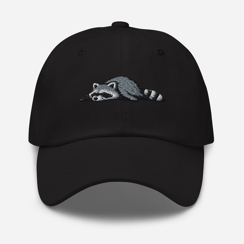 K&ouml;nnte beinhalten: Schwarze Baseballkappe mit einem gestickten Waschb&auml;ren-Design. Der Waschb&auml;r ist in Grau- und Wei&szlig;t&ouml;nen dargestellt und liegt. Die Kappe hat einen gebogenen Schirm und eine strukturierte Krone.