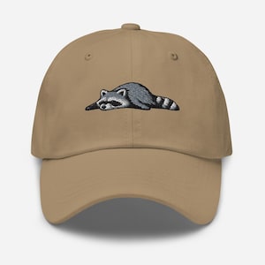 K&ouml;nnte beinhalten: Beige Baseball Cap mit einem gestickten Waschb&auml;r-Design. Der Waschb&auml;r ist in Grau und Schwarz dargestellt und liegt mit gestreiftem Schwanz. Die Kappe hat einen gebogenen Schirm und einen verstellbaren Riemen.
