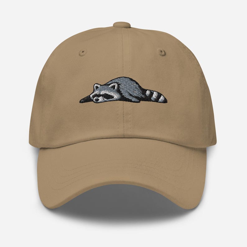 K&ouml;nnte beinhalten: Beige Baseball Cap mit einem gestickten Waschb&auml;r-Design. Der Waschb&auml;r ist in Grau und Schwarz dargestellt und liegt mit gestreiftem Schwanz. Die Kappe hat einen gebogenen Schirm und einen verstellbaren Riemen.