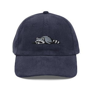 K&ouml;nnte beinhalten: Marineblaue Cord-Baseballkappe mit einem gestickten Waschb&auml;r-Design. Der Waschb&auml;r ist in Grau- und Wei&szlig;t&ouml;nen gehalten, mit einem schwarz-wei&szlig; gestreiften Schwanz. Die Kappe hat einen gebogenen Schirm und einen verstellbaren Riemen.