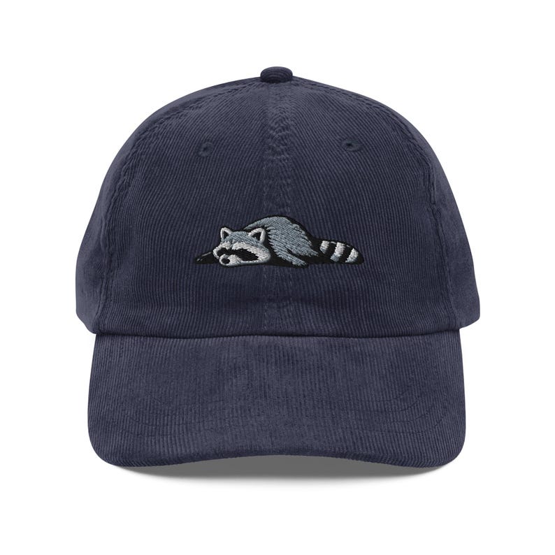 K&ouml;nnte beinhalten: Marineblaue Cord-Baseballkappe mit einem gestickten Waschb&auml;r-Design. Der Waschb&auml;r ist in Grau- und Wei&szlig;t&ouml;nen gehalten, mit einem schwarz-wei&szlig; gestreiften Schwanz. Die Kappe hat einen gebogenen Schirm und einen verstellbaren Riemen.