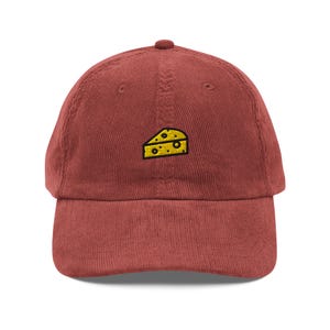 May include: A red corduroy baseball cap with a yellow embroidered cheese wedge design.