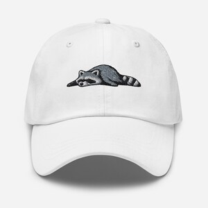 K&ouml;nnte beinhalten: Wei&szlig;e Baseballkappe mit einem gestickten Waschb&auml;ren-Design. Der Waschb&auml;r ist in Grau- und Schwarzt&ouml;nen dargestellt und liegt. Die Kappe hat einen gebogenen Schirm und einen verstellbaren Riemen auf der R&uuml;ckseite.