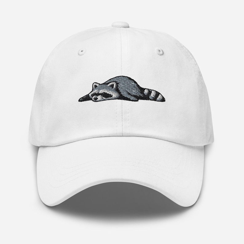 K&ouml;nnte beinhalten: Wei&szlig;e Baseballkappe mit einem gestickten Waschb&auml;ren-Design. Der Waschb&auml;r ist in Grau- und Schwarzt&ouml;nen dargestellt und liegt. Die Kappe hat einen gebogenen Schirm und einen verstellbaren Riemen auf der R&uuml;ckseite.