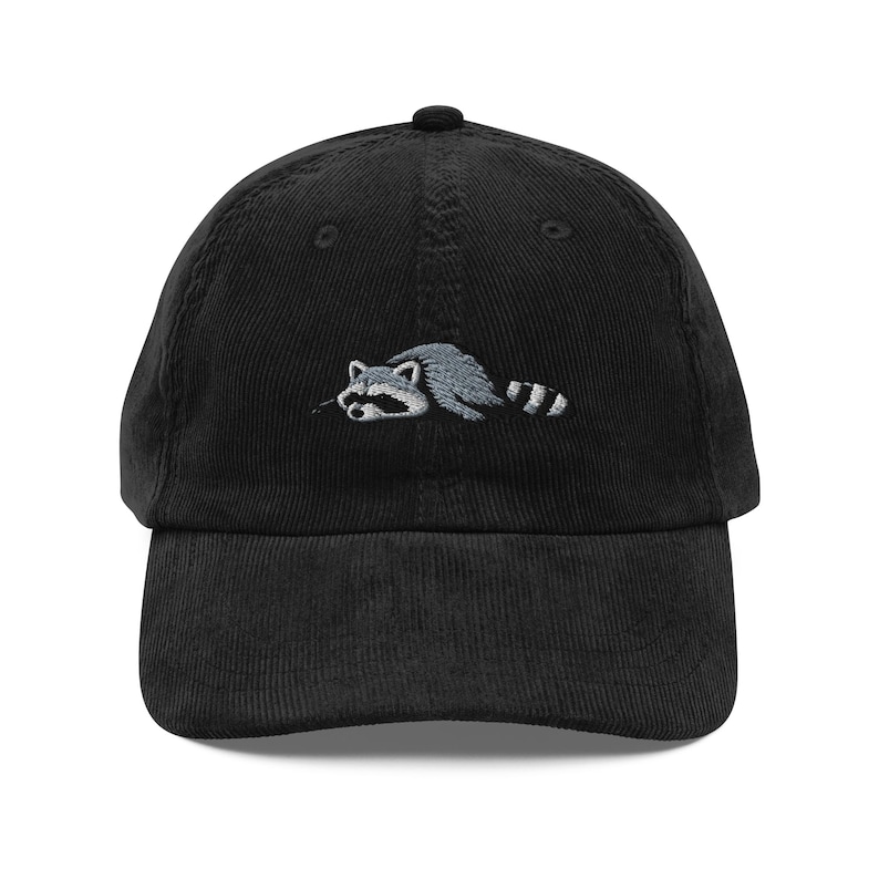 K&ouml;nnte beinhalten: Schwarze Cord-Baseballkappe mit einem gestickten Waschb&auml;r-Design in Grau und Wei&szlig;. Die Kappe hat einen gebogenen Schirm und einen verstellbaren Riemen. Ein modisches Accessoire f&uuml;r den Alltag.