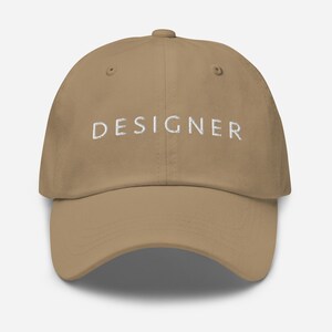 May include: A khaki dad hat with the word "DESIGNER" embroidered in white on the front.