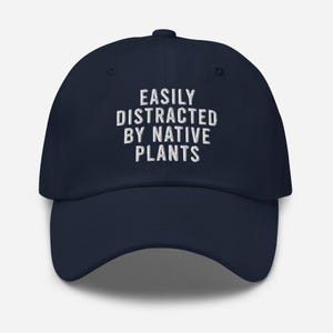 Może przedstawiać: Czapka bejsbolowa w kolorze granatowym z białym haftem z napisem "Easily Distracted By Native Plants".
