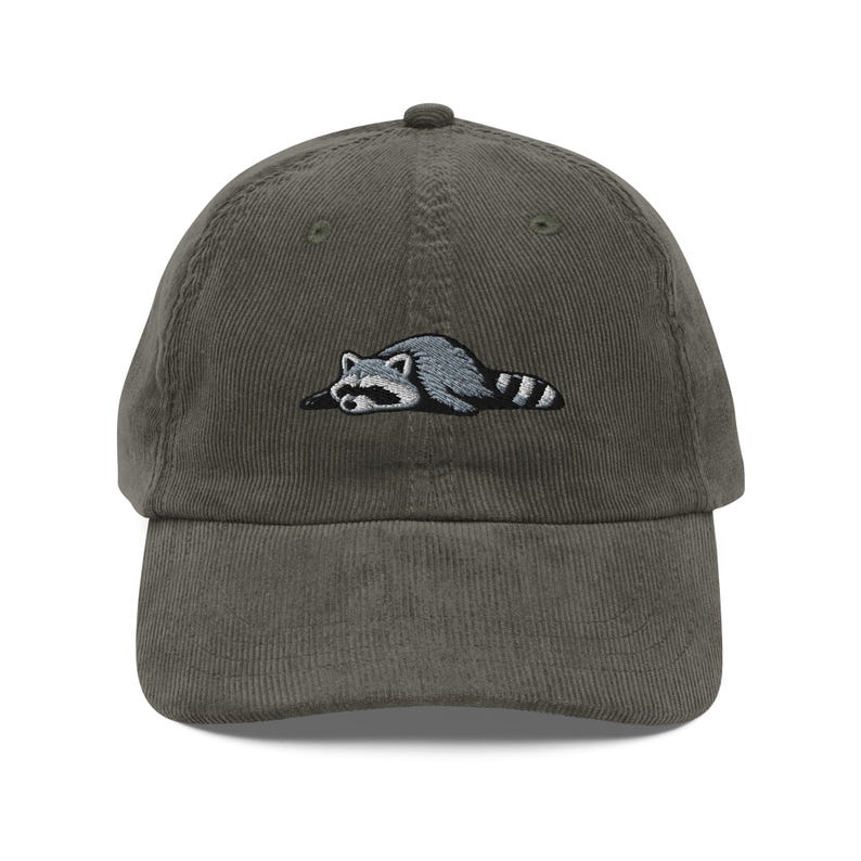 K&ouml;nnte beinhalten: Olivgr&uuml;ne Cord-Baseballkappe mit gesticktem Design eines entspannten Waschb&auml;ren in Grau- und Schwarzt&ouml;nen. Der Waschb&auml;r liegt entspannt, was dem Design der Kappe eine spielerische Note verleiht. Ein l&auml;ssiges Accessoire f&uuml;r den Alltag.