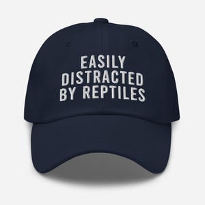 May include: A navy blue baseball cap with white embroidered text that reads "EASILY DISTRACTED BY REPTILES".