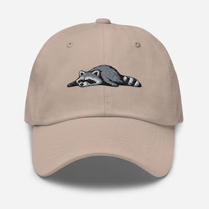 K&ouml;nnte beinhalten: Beige Baseball Cap mit einem gestickten Waschb&auml;r-Design. Der Waschb&auml;r ist in Grau und Schwarz dargestellt und liegt. Die Kappe hat einen gebogenen Schirm und einen verstellbaren Riemen auf der R&uuml;ckseite.