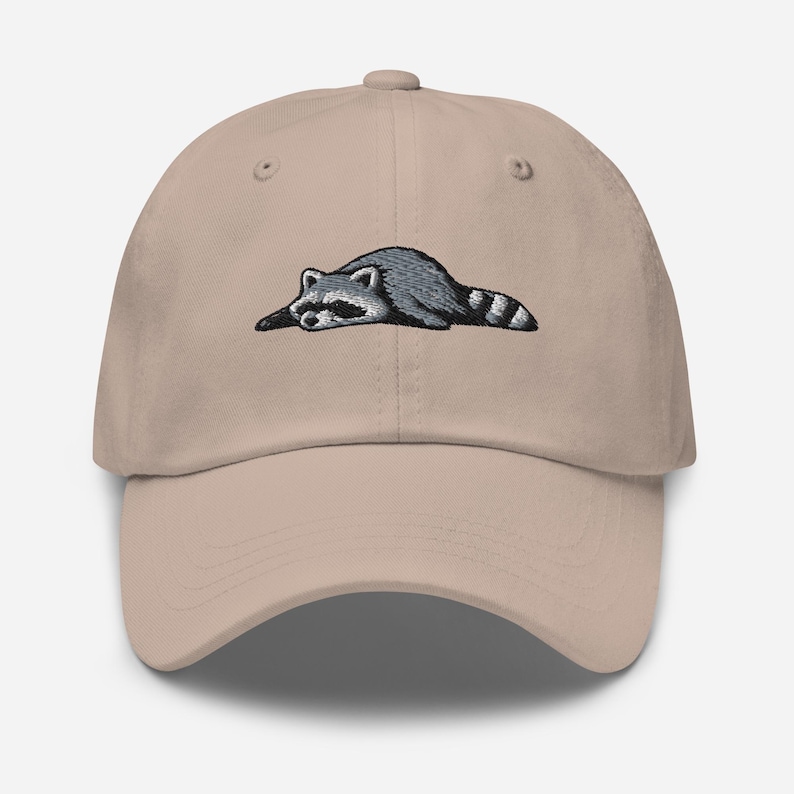 K&ouml;nnte beinhalten: Beige Baseball Cap mit einem gestickten Waschb&auml;r-Design. Der Waschb&auml;r ist in Grau und Schwarz dargestellt und liegt. Die Kappe hat einen gebogenen Schirm und einen verstellbaren Riemen auf der R&uuml;ckseite.