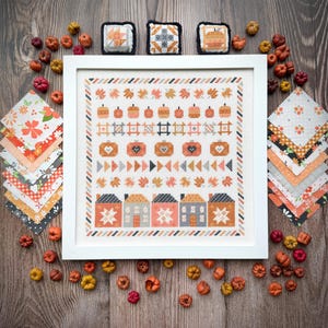 Pode incluir: Uma bordado em ponto cruz emoldurado com abóboras e casas em tons de laranja, pêssego e cinza. A obra de arte é cercada por pequenas abóboras decorativas e quadrados de tecido com estampas florais e geométricas. Três pequenas almofadas quadradas para alfinetes estão no topo.