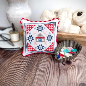May include: A small, square, white cross-stitch pillow with a red scalloped edge. The pillow features a geometric design in red, blue, and white, including a house motif. A vintage metal dish holds colorful buttons.
