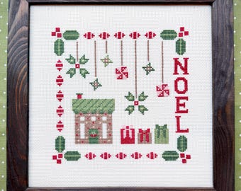 Puede incluir: Un bordado en punto de cruz enmarcado sobre tela blanca que muestra una casa, tres regalos, adornos colgantes y la palabra NOEL en rojo. El diseño utiliza hilos rojos y verdes y está dentro de un marco de madera marrón oscuro.