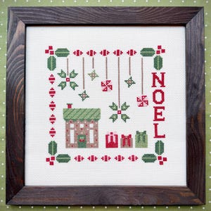 May include: A framed cross-stitch embroidery on white fabric featuring a house, three gift boxes, hanging ornaments, and the word NOEL in red. The design uses red and green thread colors and is enclosed in a dark brown wooden frame.