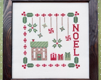 Puede incluir: Un bordado en punto de cruz enmarcado sobre tela blanca que muestra una casa, tres regalos, adornos colgantes y la palabra NOEL en rojo. El diseño utiliza hilos rojos y verdes y está dentro de un marco de madera marrón oscuro.
