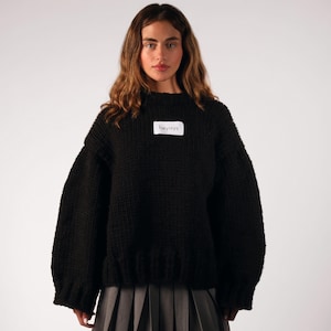 May include: A black, chunky-knit sweater with a crew neck and long sleeves. The sweater has a small white tag with the text "heyays" on the chest. The model is wearing a gray pleated skirt and black boots.