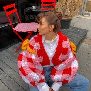Puede incluir: Un cárdigan de punto rojo y rosa a cuadros con adornos amarillos en forma de plátano en las mangas. El cárdigan es corto y tiene cuello en V.