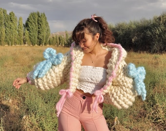 May include: A white knitted cardigan with pink and blue accents. The cardigan has a fluffy texture and features large, 3D flower embellishments on the sleeves. The model is wearing the cardigan with a white tank top and pink pants.
