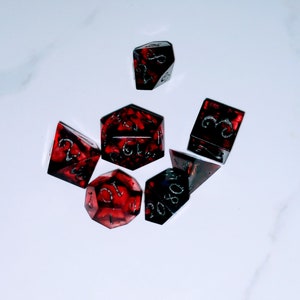 Puede incluir: Un conjunto de seis dados rojos y negros con números plateados. Los dados están hechos de un material translúcido, y los números son visibles a través de los dados.