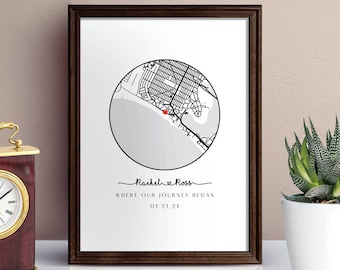 Op de afbeelding: Een ingelijst afdruk met een zwart-witte kaart van een stad met een rood hart dat een locatie markeert. De tekst onder de kaart luidt "Rachel & Ross, Where our journey began, 01.21.21". De afdruk is ingelijst in een donkerbruin frame en ligt op een wit oppervlak.