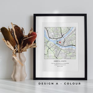 May include: A framed map print in a black frame. The map depicts a city with a river and bridges. The text "AMBER & JOSEPH" is at the bottom, along with a restaurant name, coordinates, and a date. A vase with dried flowers is on the left.