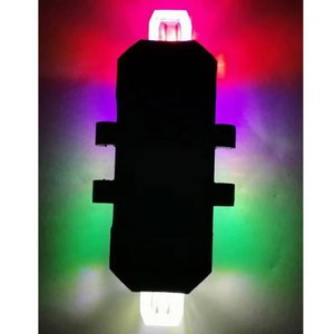 May include: A black, rectangular light with white, red, purple, and green illumination. The light has a rectangular body with two horizontal side extensions and a white light at the top and bottom.