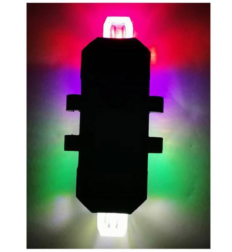 May include: A black, rectangular light with white, red, purple, and green illumination. The light has a rectangular body with two horizontal side extensions and a white light at the top and bottom.