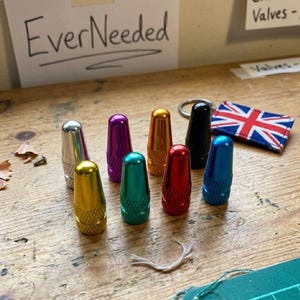 Puede incluir: Una colección de ocho coloridas tapas de vástago de válvula en plata, morado, dorado, naranja, negro, rojo, verde azulado y azul. Las tapas están dispuestas sobre una superficie de madera, con una pequeña bandera de la Union Jack y el texto "EverNeeded" al fondo.
